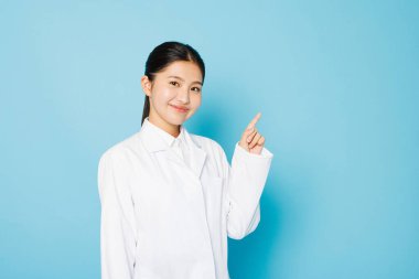 Laboratuvar önlüklü mutlu öğrencinin portresi mavi arka plandaki kameraya parmak işareti yapıyor ve gülümsüyor.