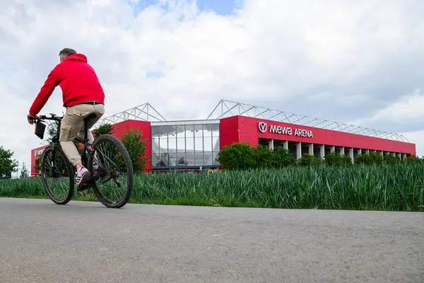 MAINZ, GERMANY - 17 Mayıs 2025: Bir adam MeWA ArENA 'nın önündeki çiçek açan kır çiçeği tarlasının önünden bisikletle geçiyor, renkli çiçek açan kır çiçekleri tarlasının arkasında görülüyor. Bundesliga eşleşmesi öncesinde manzaralı atmosfer oluşturuluyor.