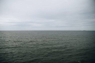 Karanlık, dalgalı bir denizin bulutlu bir gökyüzünün altında geniş bir manzarası. Ufuk uzaklıktan zar zor görünüyor. Minimalist ve kasvetli bir deniz manzarası yaratıyor..
