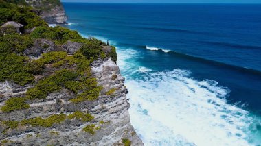 Kızgın okyanusun üzerindeki yüksek kayalık uçurum. Uçurumun kenarında siyah elbiseli genç bir kadın duruyor. Hava fotoğrafı, yan görüş. Hayalperest, yalnız ve düşünceli olma kavramı..