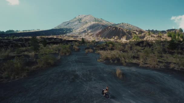バリ島のバルタ火山にモペットに乗った若い女性の空中撮影 黒い溶岩の火山への道 山と空の景色 極端な状態でモペットに乗る — ストック動画