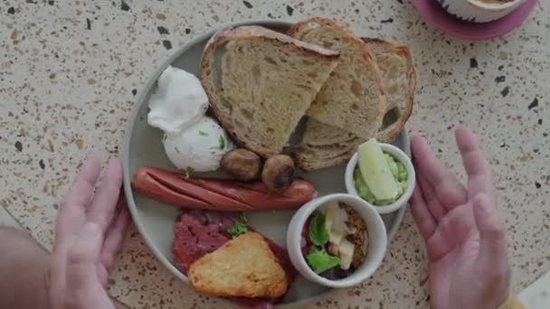 イギリスの伝統的な朝食は男性が取ったプレートです 卵ベネディクト 揚げソーセージ ベーコン トーストを含む伝統的な朝食のプレートを取る男性の手のクローズショット — ストック動画