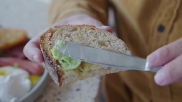 熟した緑色のアボカドをマッシュした全粒粉のトーストパンのクローズアップショットは ナイフでパンに広がります ケトダイエットやビーガンに適した便利で健康的な朝食 — ストック動画