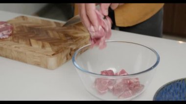 Genç bir adam ahşap bir kesme tahtasına çiğ et kesiyor ve et parçalarını şeffaf bir tabağa döküyor. Mutfakta ağır çekimde yemek pişirmek..