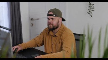 Evden çalışan, günlük giysiler içinde sakallı bir adamın ön görüntüsü. Uzak bir serbest iş yeri. Bir adam online bir işte çalışıyor. Bilgisayarda daktilo ediyorum, konsantre oluyorum..