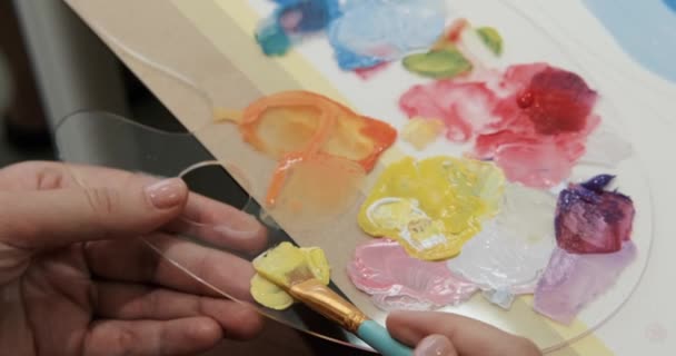 パレットにアクリルペイントを混ぜる若い女性の手のクローズアップ 初心者のためのレッスン 絵画のための色を学びます マルチカラーの塗料がブラシと混合されています — ストック動画