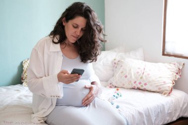 Koca göbekli hamile bir kadın yatağa oturur ve elinde bir telefon tutar. Hamile kadınlar için mobil başvuru formu. Bir kadın mobil bir uygulamayla doğuma ne kadar zaman kaldığına bakar..