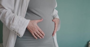 Gündelik giysiler içinde koca göbekli hamile bir kadın odasında karnını okşuyor. Sakin annelik ve doğum. Hamilelik gelişimi için jinekolojik izleme.
