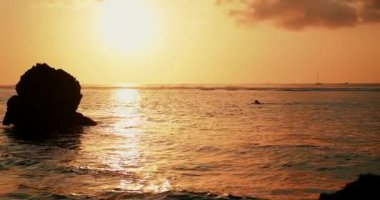 Gün batımında sörfçünün sörf tahtasındaki görüntüsü. Altın saat, batan güneş gökyüzünü ve suyun yüzeyini altın ışıkla boyar. Bir sörfçü okyanustan sahile döner..