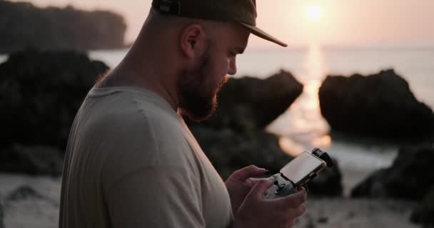 男は日没時に海の近くのビーチでドローンを飛ばし サーファーのビデオを撮る 最新技術 無人制御ドローン コントロールパネル 接続された携帯電話 — ストック動画