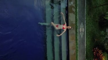 Kırmızı bikinili yüzme havuzunda yatan genç bir kadının dikey videosu. Uzun zamandır beklenen Bali tropikal adasında bir tatil. Seyahat ve barış kavramı.