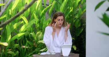 Güzel, orta yaşlı bir kadının beyaz bir bornoz giyip gözlerinin altına yaşlanma karşıtı krem sürmesi ve aynaya bakması. Yaşlanan yüz derisine dikkat et.