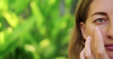 Orta yaşlı güzel bir kadının yüzüne yakın dur. Bir kadın gözlerinin altına krem sürer masaj hareketleriyle, yaşlanma karşıtı cilt bakımıyla. Yeşil yapraklı doğal arkaplan, metin için boş alan.
