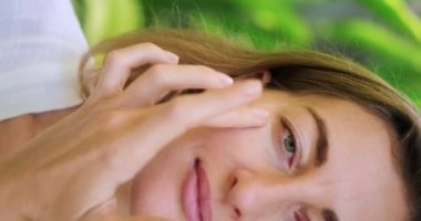 Orta yaşlı bir kadının yüzünün dikey yakın çekim videosu. Bir kadın gözlerinin altına krem sürer masaj hareketleriyle, yaşlanma karşıtı cilt bakımıyla. Yeşil yapraklı doğal arkaplan, boş alan
