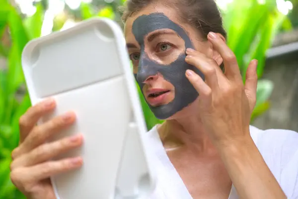 A middle aged woman applies a gray clay mask to her face and looks in the mirror. A woman in a courtyard against a backdrop of green foliage in the tropics applies a clay mask to cleanse her skin.