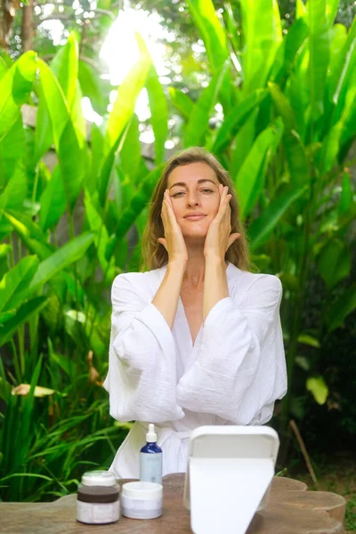 Medium shot of a beautiful middle-aged woman sitting in a garden wearing a white robe and applying anti-aging cream under her eyes. Care for aging facial skin.