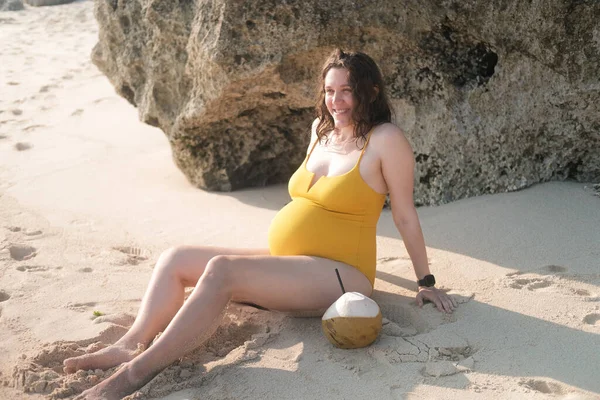 A happy pregnant woman with a big belly sits on the beach near the ocean. Third trimester of pregnancy in the tropics on vacation. Pregnancy and preparation for childbirth, relaxation on the beach.