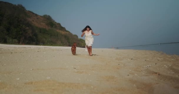 一个身穿连衣裙的年轻女子和一只金黄色的猎犬沿着海滩奔向摄像机的前视图 在海滩上和狗玩积极的游戏 狗是人类最好的朋友 — 图库视频影像