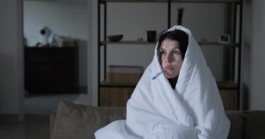 Soğuk algınlığı olan bir kadın başını battaniyeye sararken termometreyle ısısını ölçer. Evde yüksek sıcaklık, tedavi ve sağlık hizmetlerinden kaynaklanan titreme ve ateş.
