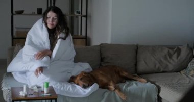 Genç bir kadın nezle olur, oturma odasındaki kanepede oturur, battaniyeye sarınır, yanında bir köpek yatar. Bir kadın burnunu mendile sümkürdü. Sümük ve öksürük, soğuk algınlığı.