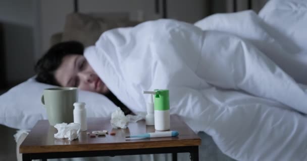 一位生病的年轻女子躺在沙发上 面前的一条毯子下 一张附有药物的床头柜 一个女人伸手拿温度计量体温 发烧及体温过高 — 图库视频影像