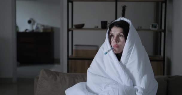 寒い女性は毛布に頭を包んで温度計で体温を測定する 自宅での高温 医療からのチルと発熱 — ストック動画
