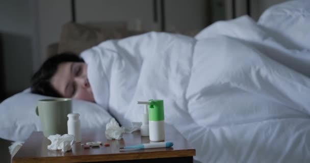 一位患感冒的年轻女子躺在客厅的沙发上 面前是一张床头柜 桌上放着药品和餐巾 一个女人得了高烧高烧 测量温度 — 图库视频影像