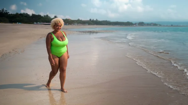 A full length, plus size African American woman with curly hair in a light green swimsuit walks along the beach near the ocean. The concept of body acceptance and body positivity. Banner, free space.
