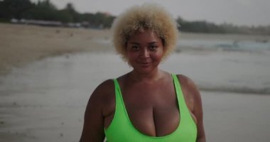 Sahilde yürüyen ve flört eden iri yarı bir Afro-Amerikan kadın. Mayo giymiş iri göğüslü güzel bir Afro-Amerikalı kadın kameraya doğru yürüyor..