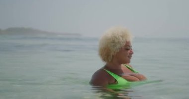 Sarı kıvırcık saçlı, yeşil mayo giyen, genç ve iri yarı Afro-Amerikan kadın okyanusta yüzüyor ve tatilinin tadını çıkarıyor. Vücut pozitifliği ve kendini kabul etme kavramı..