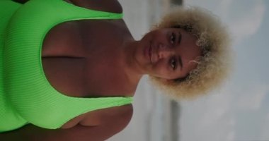 Sahilde yürüyen ve flört eden iri yarı Afro-Amerikan bir kadının dikey videosu. Mayo giymiş iri göğüslü güzel bir Afro-Amerikalı kadın kameraya doğru yürüyor..