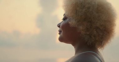 Sarışın, kıvırcık saçlı, okyanus kıyısında güneşin doğuşunu seyreden ve gülümseyen Afro-Amerikan bir kadının yüzünün yan görüntüsü. Sahilde güneşin doğuşunun tadını çıkarıyorum..