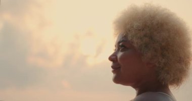 Güneş doğarken kumsalda durup gülümseyen beyaz kıvırcık saçlı Afro-Amerikan bir kadının yüz resmi. Şafakta güneş ışığı, plajda altın saat. Güneşin altında portre.