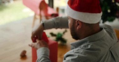 Noel Baba şapkası takan, Noel Baba şapkalı ve yeni yıl hediyelerini salonda paketleyen sakallı bir adamın yakın plan fotoğrafı. Noel kutlaması ve hediye paketleme hazırlığı.
