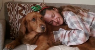 Ren geyiği boynuzlarıyla Noel kostümü giymiş genç bir kadın kanepeye uzanmış köpeğiyle onu okşuyor. Yeni yılı ve Noel 'i bir köpekle kutlamak.