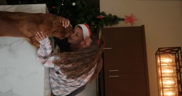 一对恋爱中的年轻夫妇在厨房里庆祝圣诞节的垂直录像 背景是一棵圣诞树 爱抚着他们的金毛猎犬 假日的气氛 — 图库视频影像