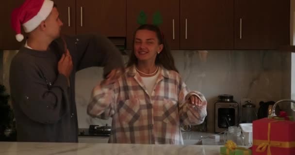 一个戴着圣诞礼帽的男人在厨房为他的妻子唱了一首歌 圣诞节前夕 一对恋爱中的年轻夫妇在圣诞树旁的厨房里跳着浪漫的舞 — 图库视频影像