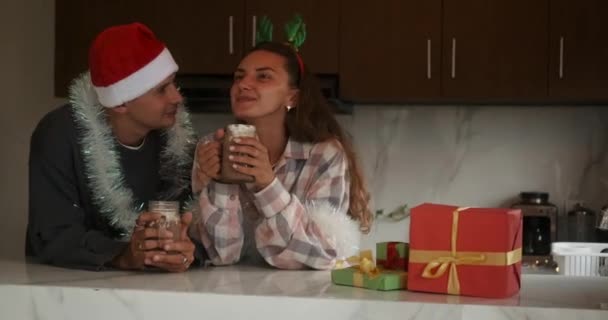 一对年轻夫妇在厨房的新公寓里 用棉花糖喝可可 一个头戴圣诞礼帽的男人和一个年轻女人在窃窃私语和大笑 圣诞气氛 — 图库视频影像