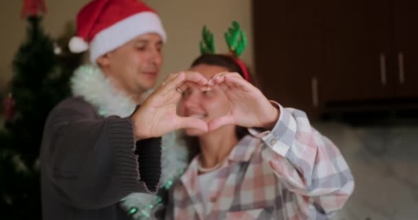 一对恋人在圣诞树的背景下 在他们的微笑中 展示了一个用手指做成的心形符号 一个戴着圣诞礼帽的男人吻了一个年轻的女人 圣诞之心 家庭传统 — 图库视频影像