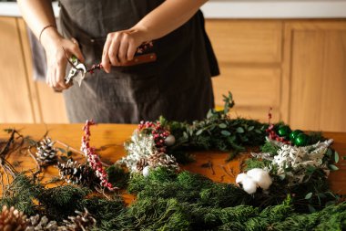 Elinde tel makası olan genç bir kadın Noel çelengi yapmak için ladin ağacından dallar kesiyor. Noel ve Yeni Yıl için dekorasyon üzerine atölye.