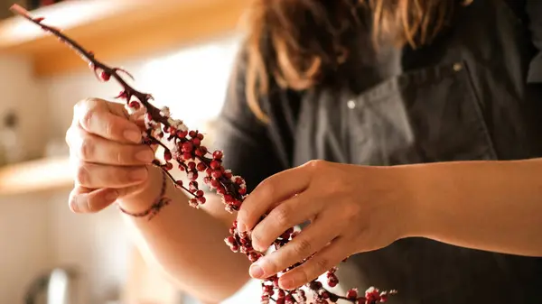 A branch with red decorative berries is in the hands of a young woman who makes a Christmas wreath and decorated it. Preparing and decorating your home for the New Year and Christmas.