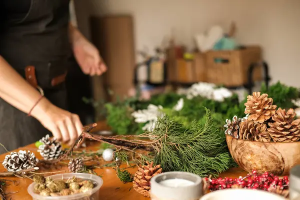 Noel çelengi hazırlama atölyesi. Genç bir kadın ahşap bir masanın üzerinde köknar dallarından bir çelenk topluyor. DIY dekorasyonları. Masadaki çelenk için süslemeler var: kozalaklar ve kurdeleler..