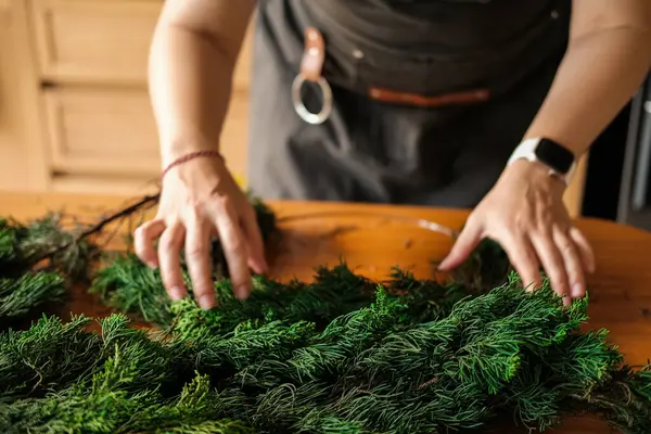 Tahta bir masadaki köknar dallarından Noel çelengi toplayan genç bir kadının yakın çekimi. Yeni yıl ve Noel için süslemeler hazırlanıyor.