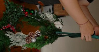 Genç bir kadının evdeki mutfağında yaptığı Noel çelengine kurdele bağlarken çekilmiş dikey videosu. Evde Noel süsü yapmak için özel ders. Çelenk koni ve toplarla süslenmiş..