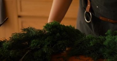 Tahta bir masadaki köknar dallarından Noel çelengi toplayan genç bir kadının yakın çekimi. Yeni yıl ve Noel için süslemeler hazırlanıyor.