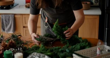 Önlüklü genç bir kadın köknar dallarından Noel çelengi yapıyor. Yeni yıl için dekorasyonlar masaya saçılmıştır: mumlar, kozalaklar, Noel ağacı topları ve bir yıldız..