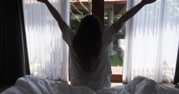 从一个坐在床上 靠着窗户伸展着的年轻女子的背后看去 夏天醒来的概念 在窗户的花园里 有绿叶的树木 — 图库视频影像
