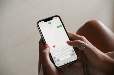 Telefonda kalp şeklinde bir aşk ve emoji beyanı içeren bir mesaj yazan genç bir kadının ellerinin yakın çekimi. Sevgililer Günü Sevdiğine mesaj at.