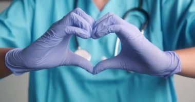 Tıbbi üniformalı ve boynunda steteskop olan mavi eldivenli bir kadın doktora orta boy iğne. Elleriyle bir kalp sembolü gösteriyor. Sevgililer Günü doktoru bir tıp merkezinde. Kardiyolog.
