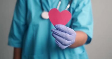 Tıbbi üniformalı, mavi eldivenli ve boynunda steteskop olan kadın bir doktor ellerinde kağıttan yapılmış kırmızı bir kalp tutuyor. Doktorda sevgililer günü. Kardiyoloji konsepti.